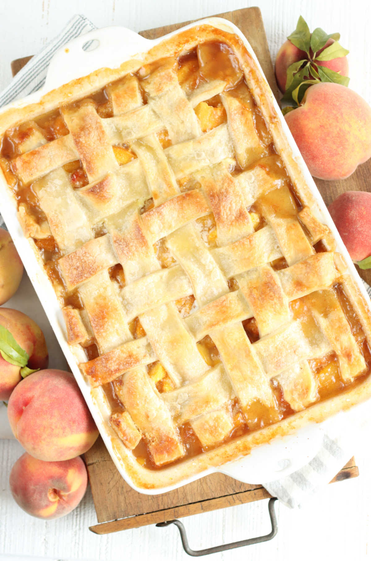 Cobbler with peaches and lattice pie crust in white rectangle baking dish on wooden cutting board, fresh peaches around.