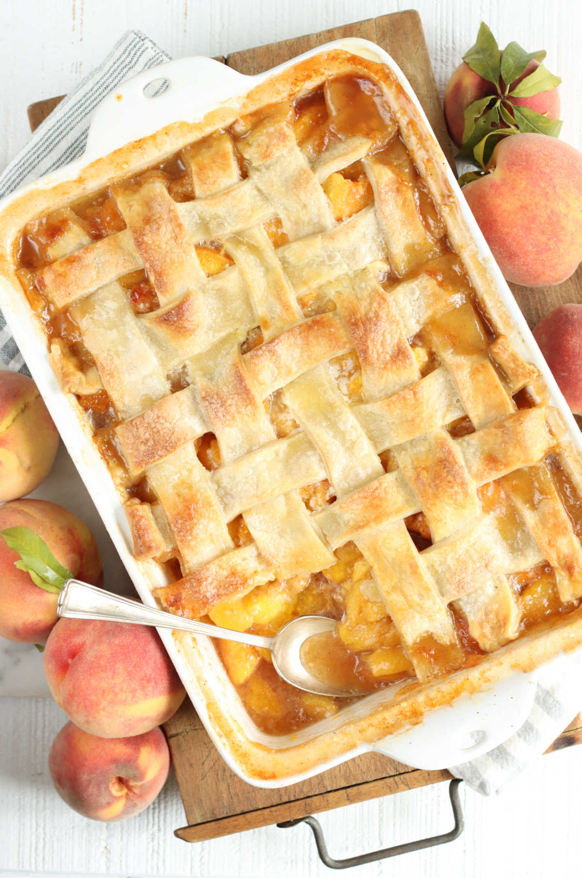 Peach cobbler with pie crust lattice weaved in white rectangle baking dish on wooden cutting board.