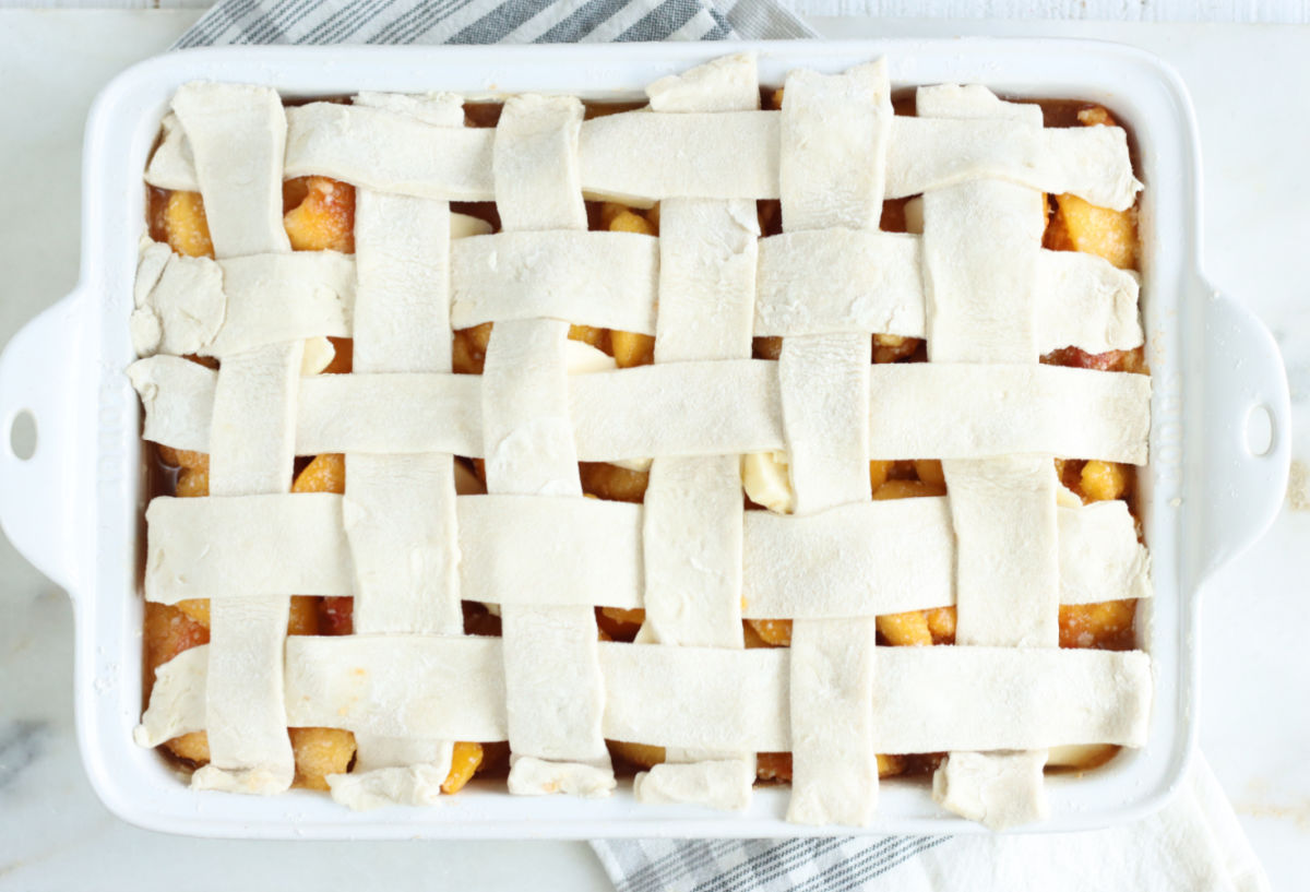 Lattice pie crust on peach cobbler in white baking dish on white marble.