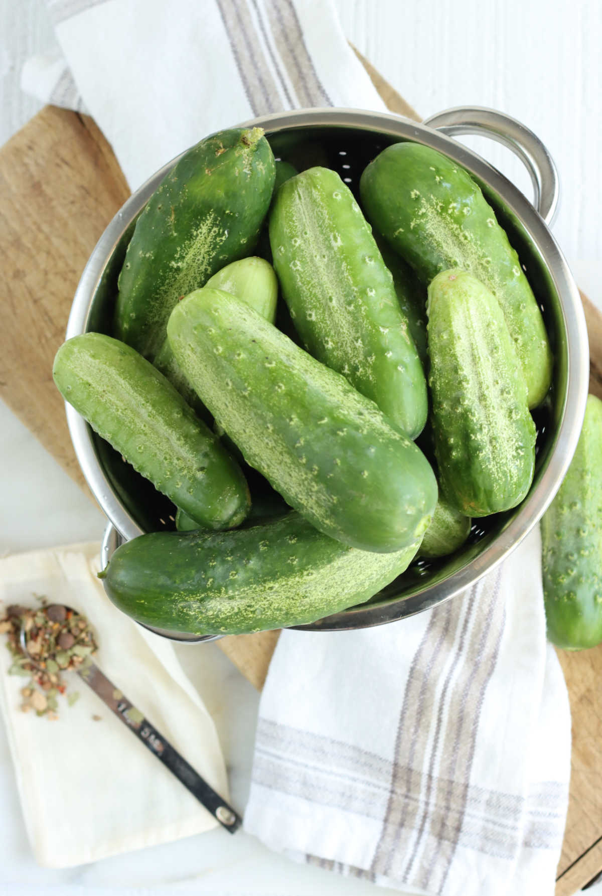 Stainless steel strainer with pickling cucumbers on wooden cutting board, teaspoon of pickling spice.