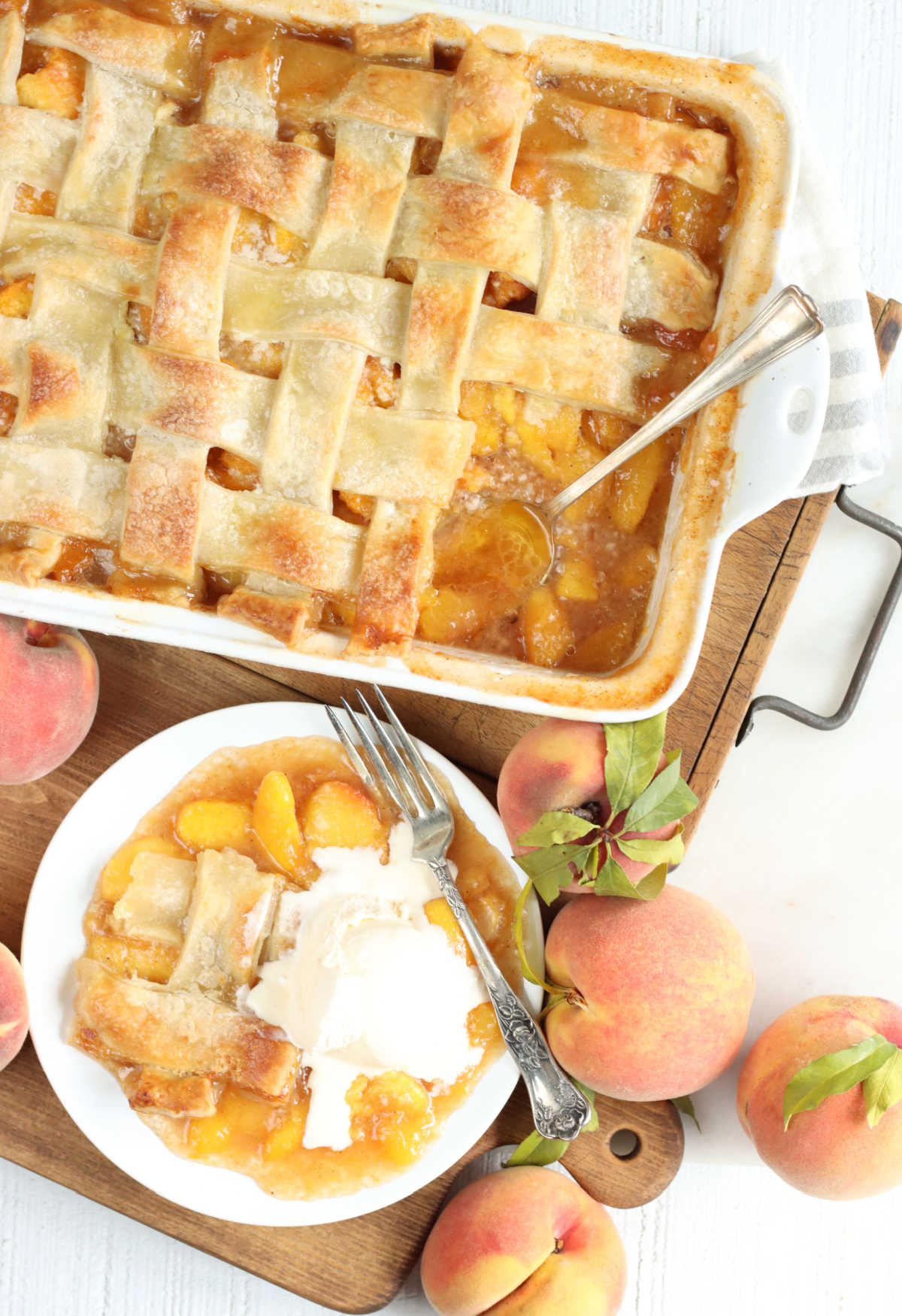 Peach cobbler in white baking dish, white plate with piece of cobbler and fork, fresh peaches around.