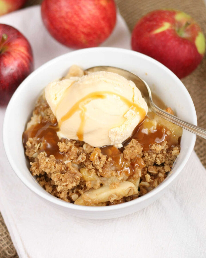 Apple crumble topped with vanilla ice cream, caramel sauce, spoon in small white bowl.