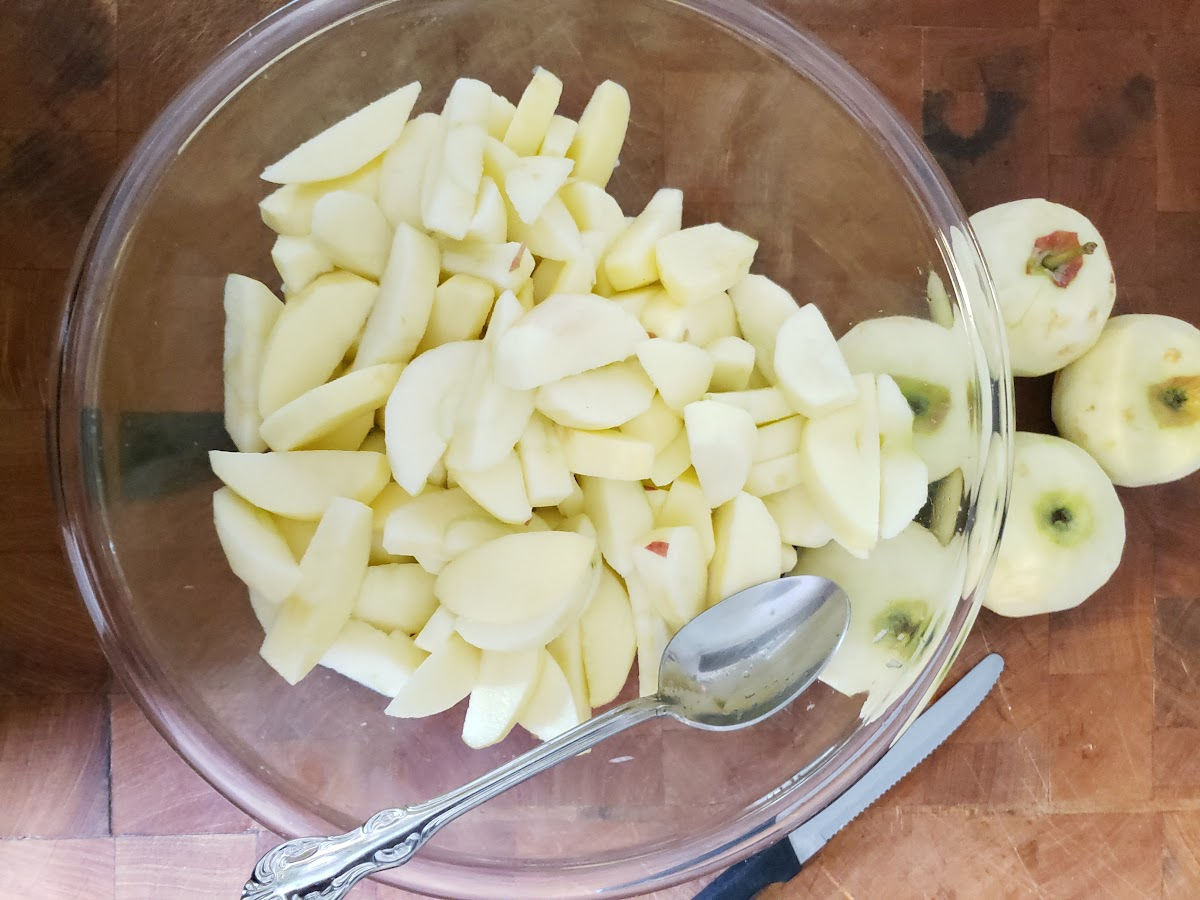 Peeled apple slices in clear glass bowl on butcher block, spoon in bowl.