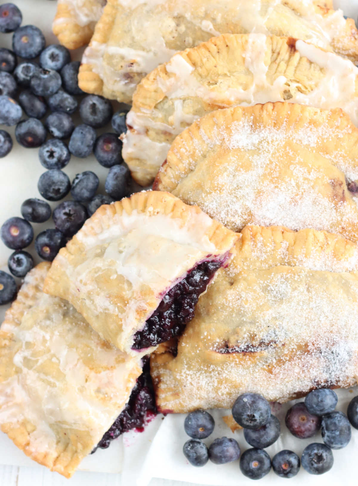 Blueberry hand pies on white marble, one cut in half, fresh blueberries around.