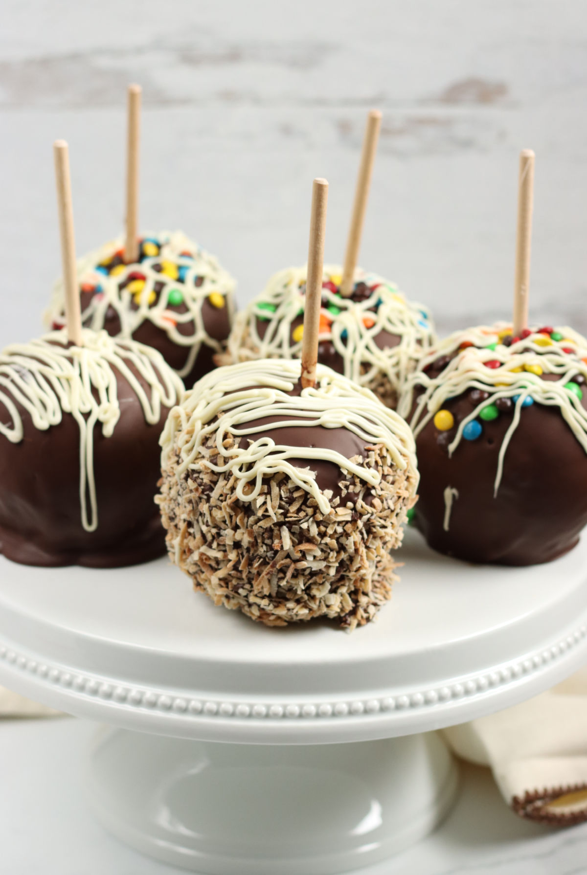 Caramel chocolate apples rolled in candies on white footed cake dish.