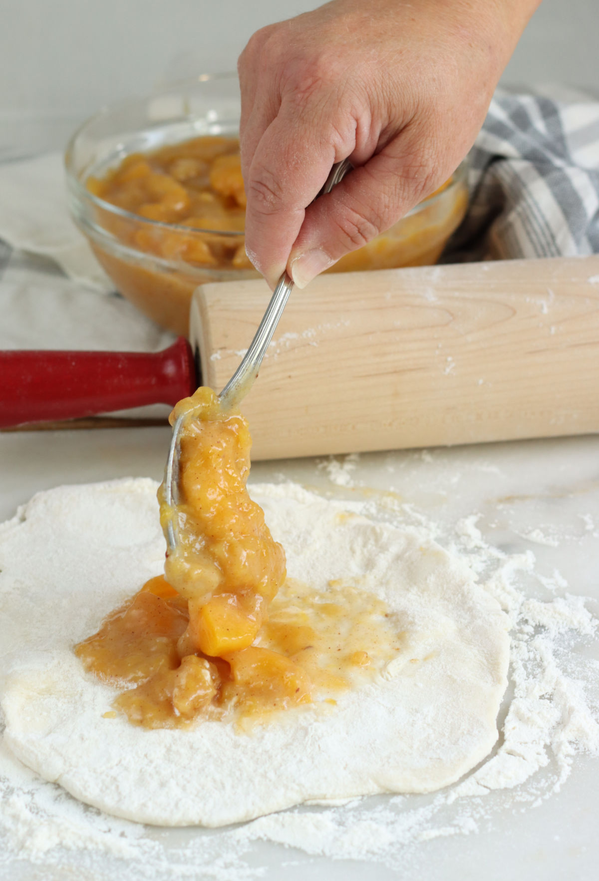 Filling hand pie with peach pie filling on white marble, red handle wooden rolling pin in background.