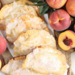 Peach hand pies on burlap, fresh peaches and halves around.
