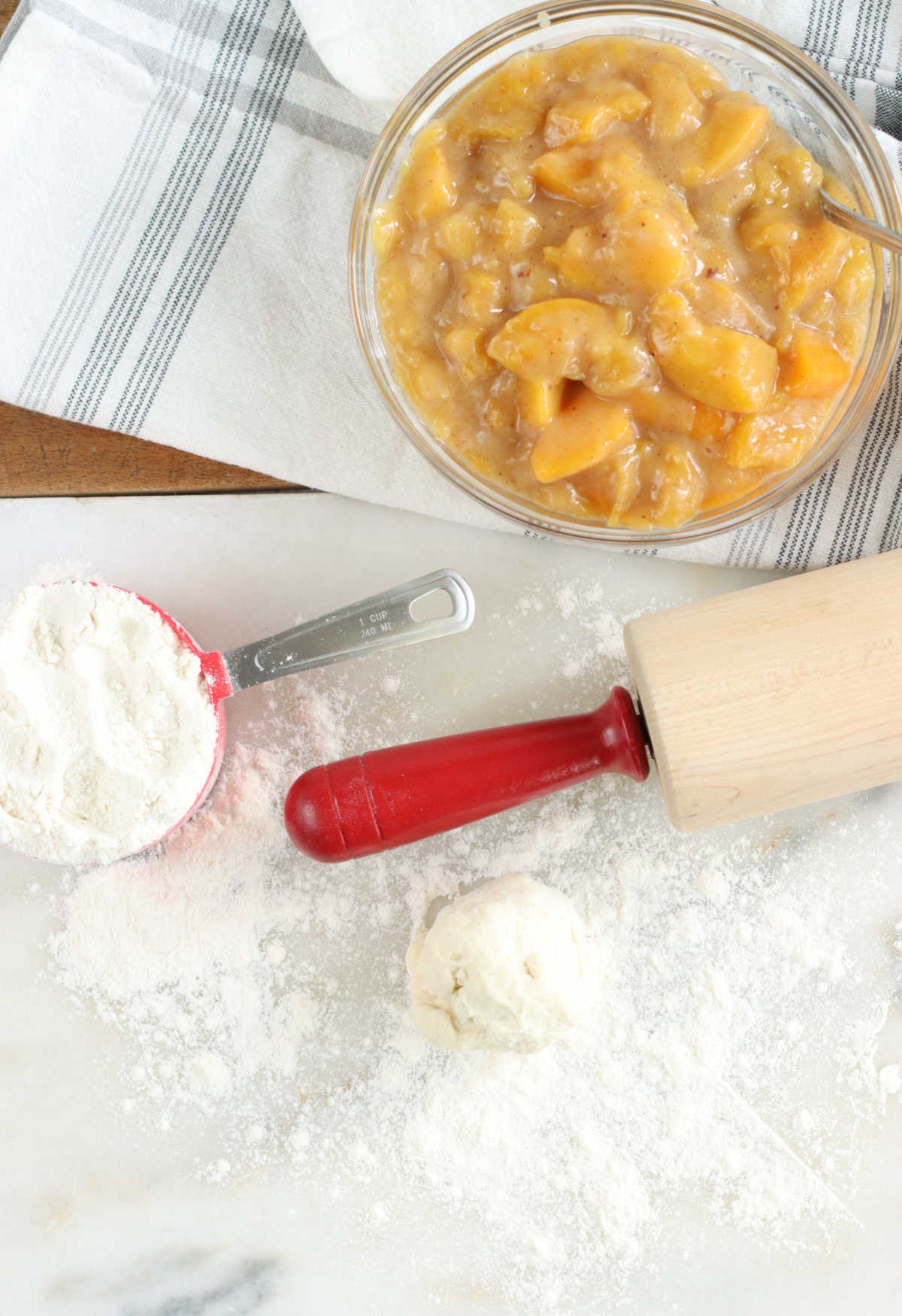 Pie crust round, peach pie filling in clear glass bowl, wooden rolling pin with red handles on white marble.