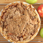 Apple pie with crumb topping on butcher block, loose apples around.