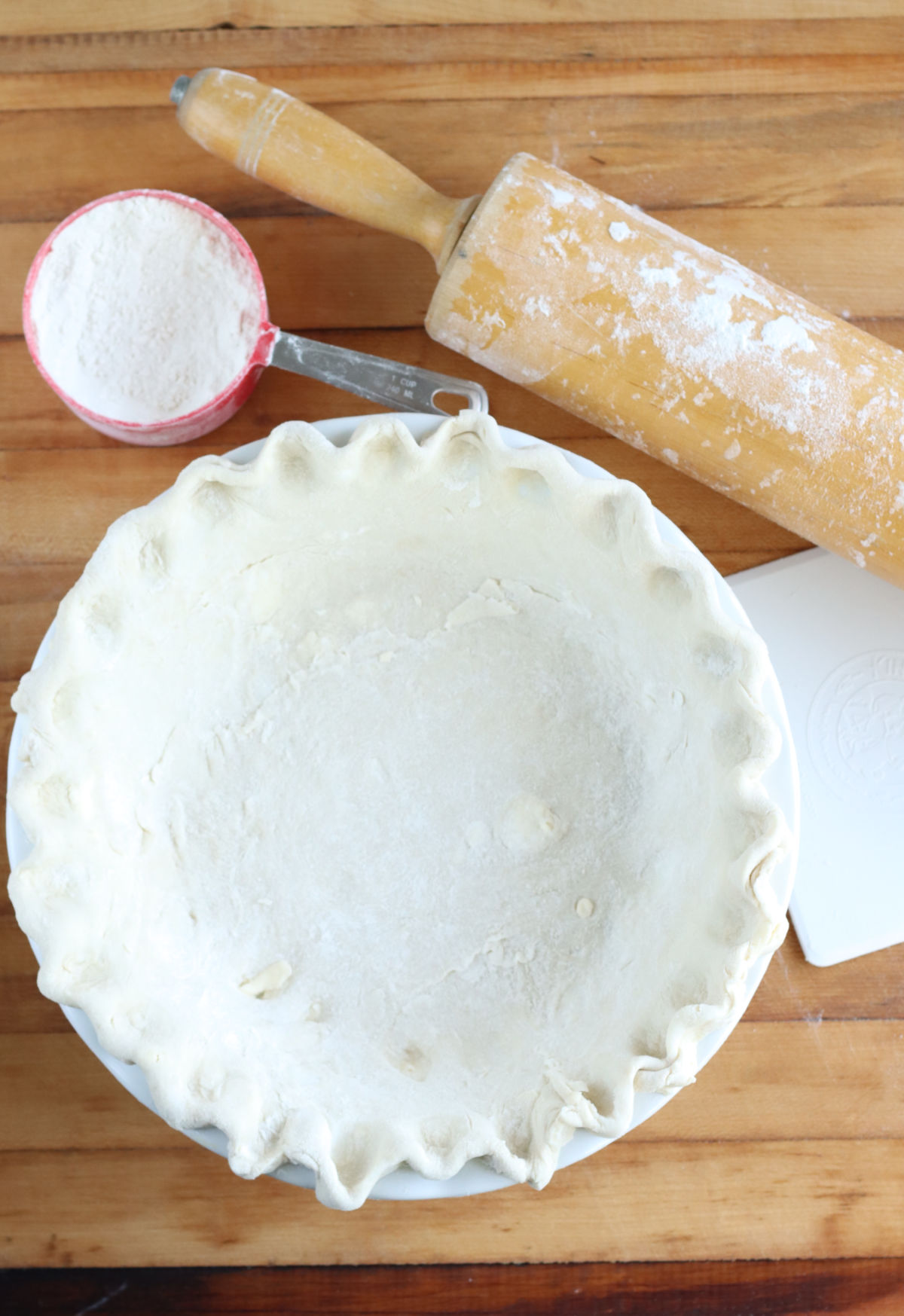 Unbaked pie crust in white pie dish on butcher block, wooden rolling pin, red measuring cup with flour.