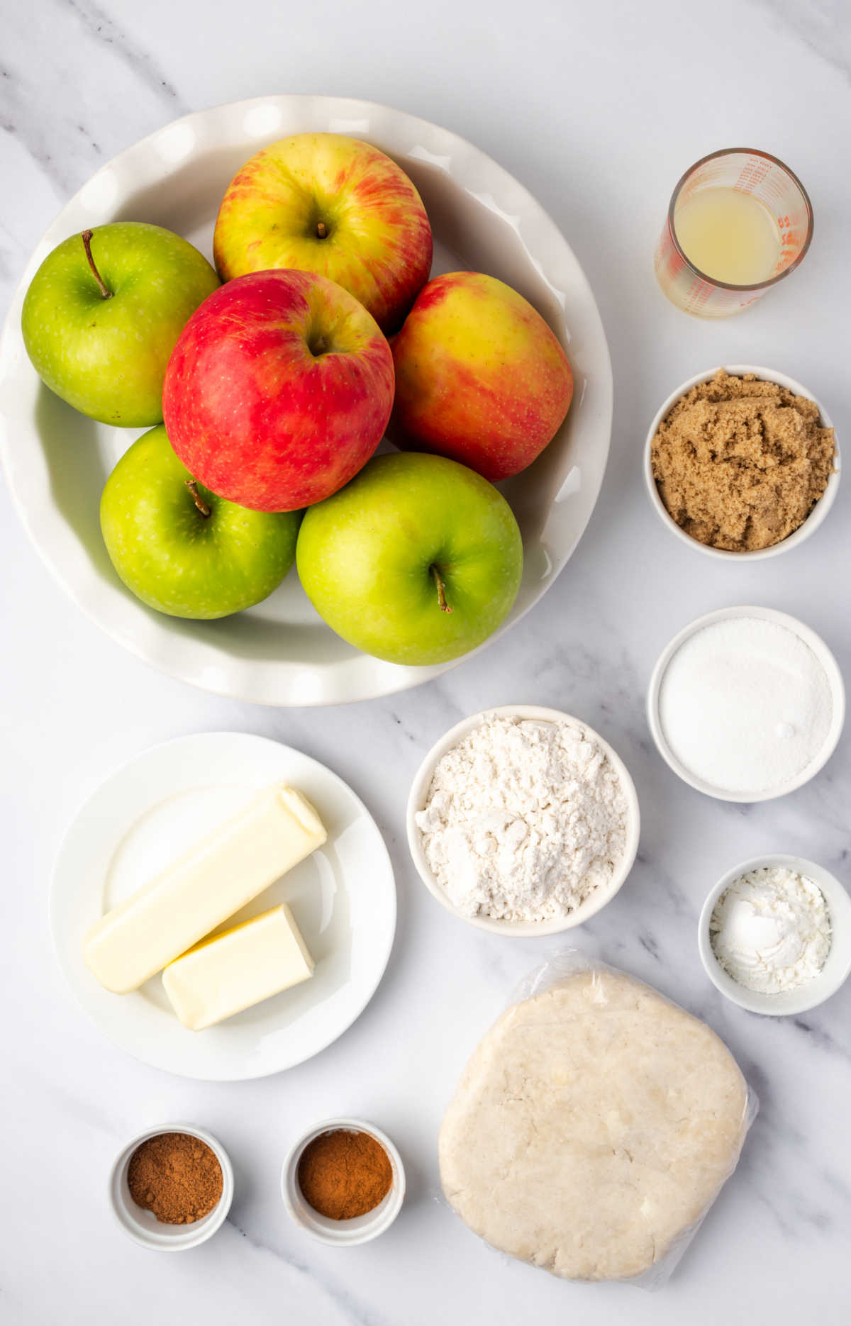 Ingredients in small white bowls for apple pie, apples in white pie dish.