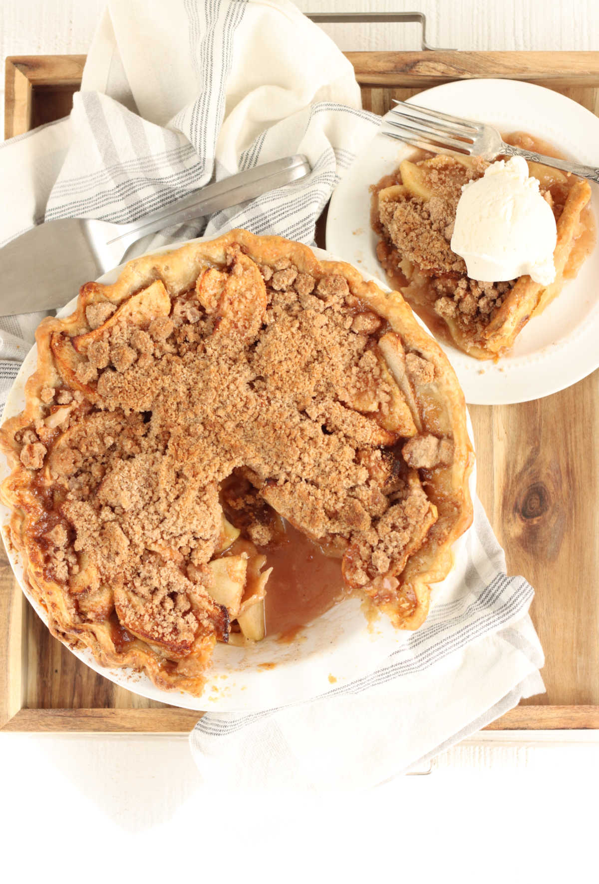 Apple pie with crumb topping, slice missing in wooden box, slice on small white plate with fork.
