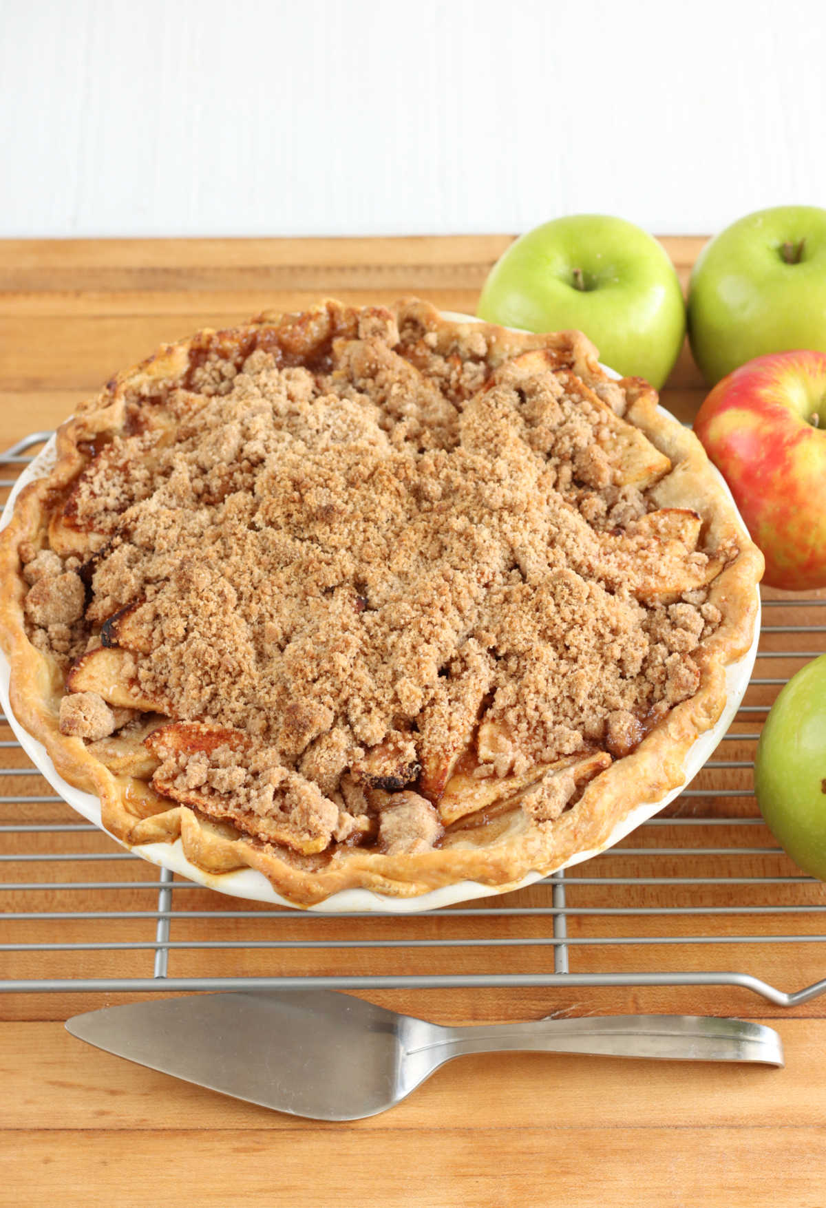 Apple pie with crumb topping on metal cooling rack on butcher block, pie cutter in front.