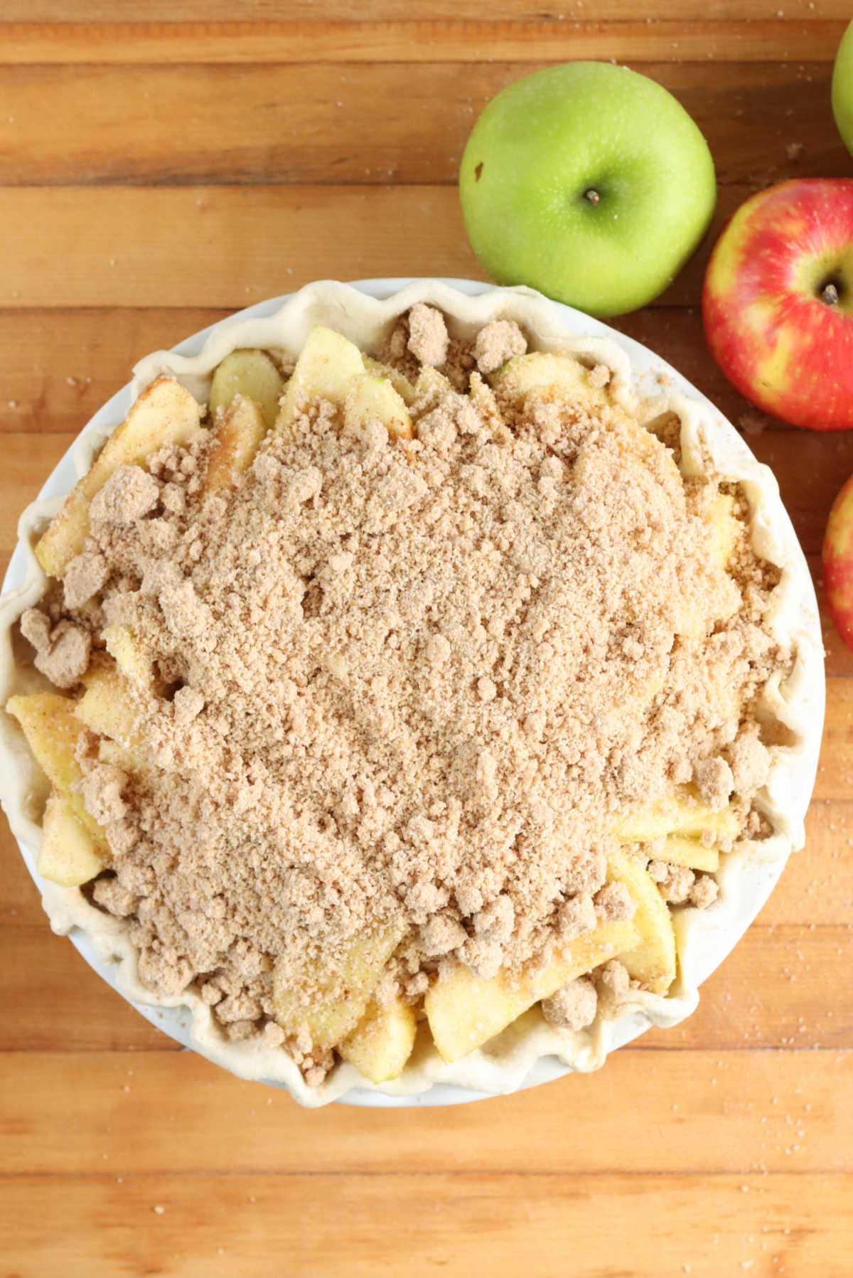 Unbaked apple pie with crumb topping on butcher block, apples around.