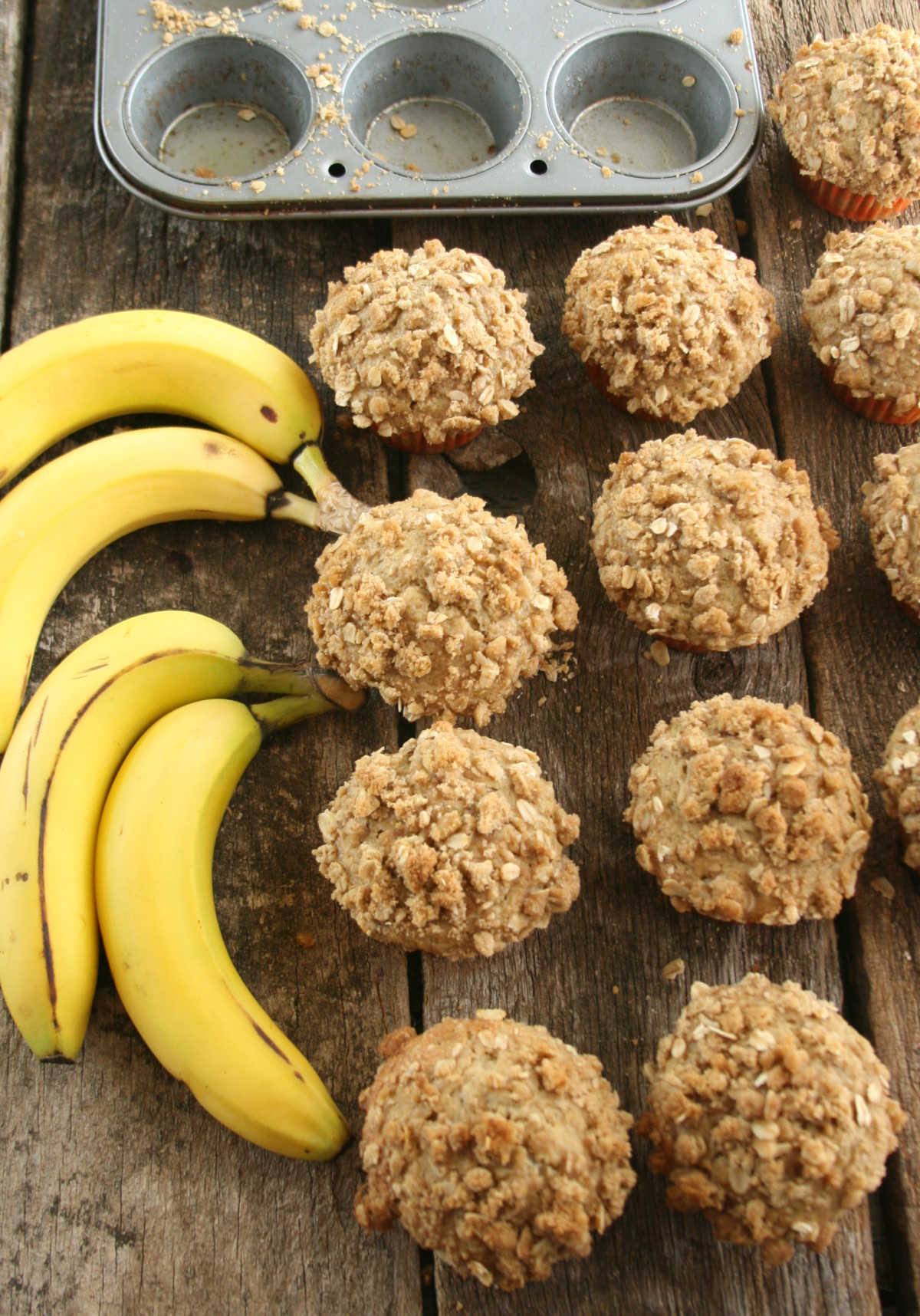 Banana muffins with crumb topping on reclaimed boards, muffin tin behind, ripe bananas to side.