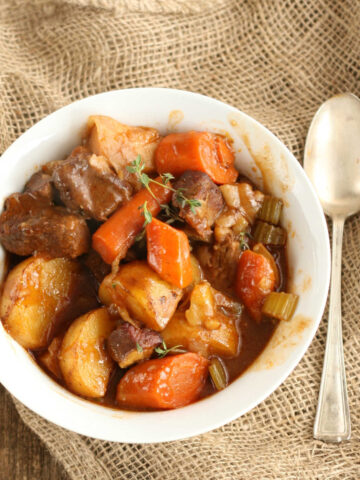 Beef stew with carrots, potatoes, celery in small white bowl, spoon on right.