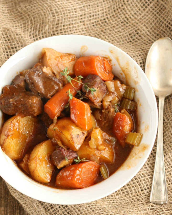Beef stew with carrots, potatoes, celery in small white bowl, spoon on right.