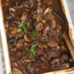 Stew beef and brown gravy in white rectangle baking dish, wooden spoon.