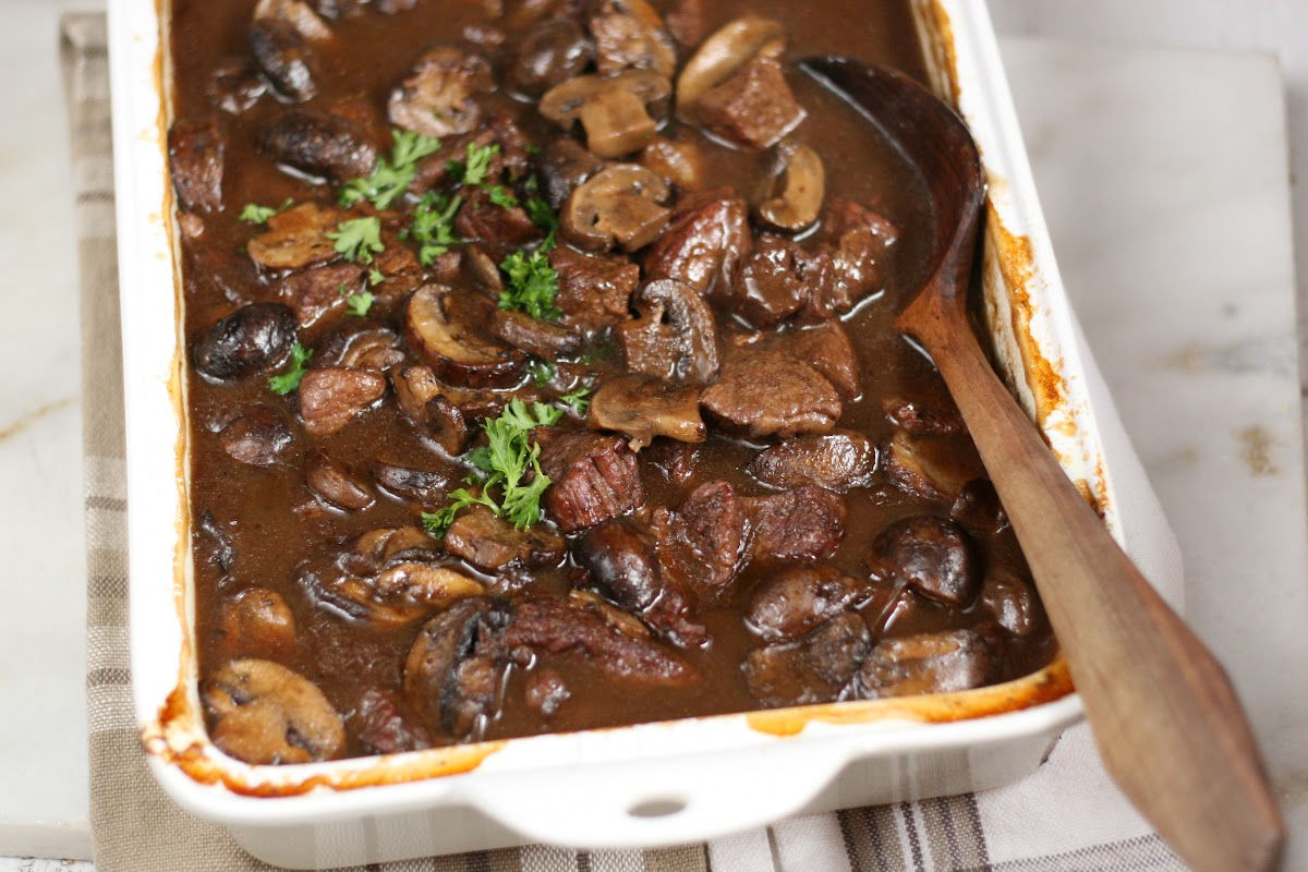 Stew beef in mushroom gravy in white rectangle baking dish, wooden spoon in right of dish.