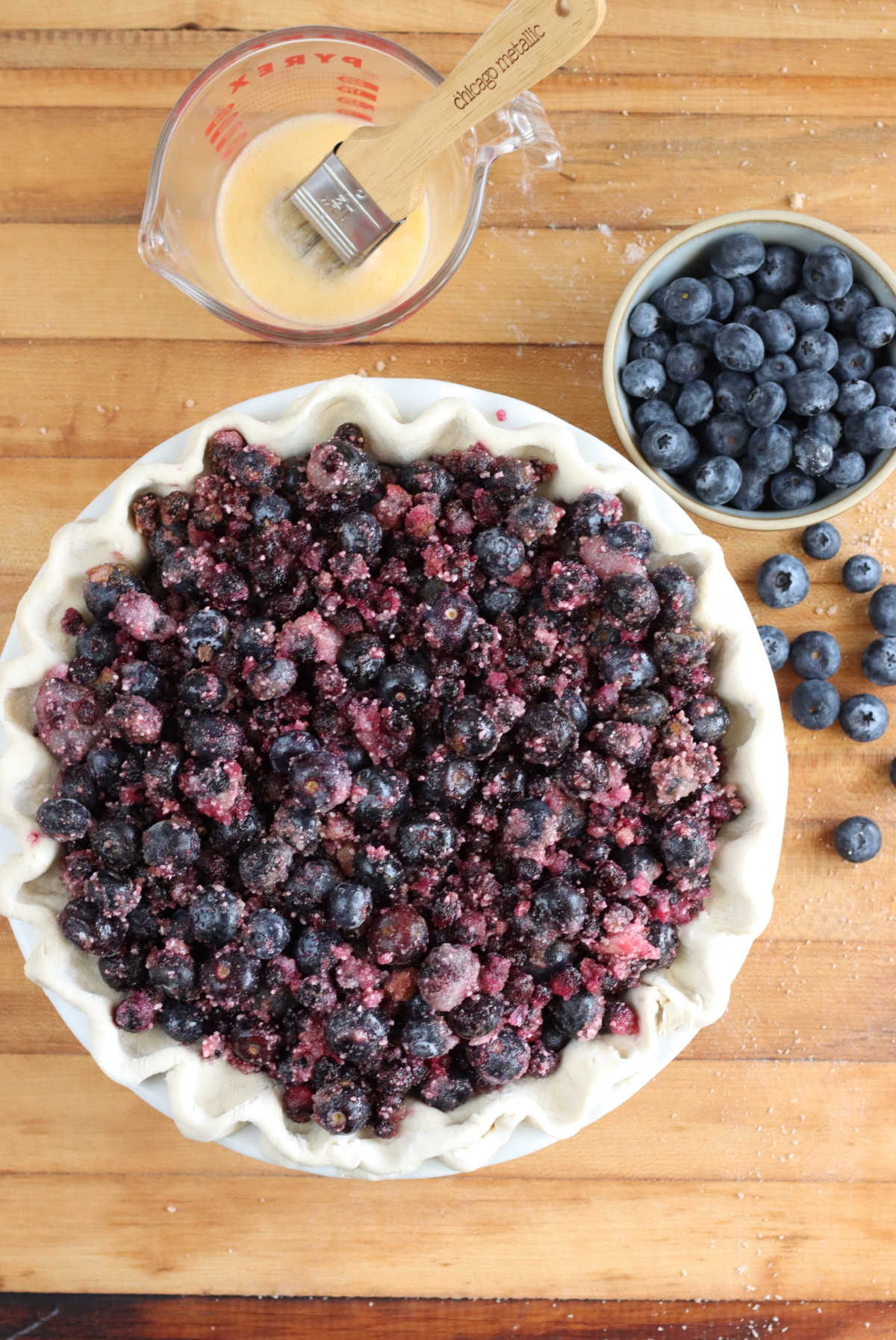 Blueberry pie filling in unbaked pie shell on butcher block, egg wash and pastry brush in glass measuring cup.