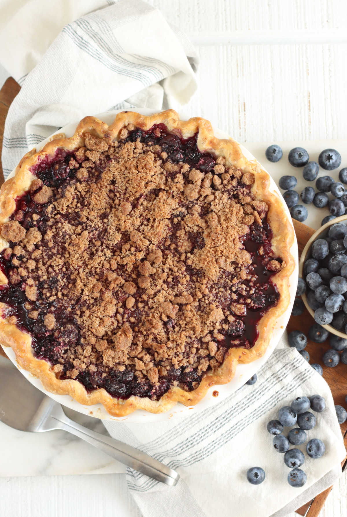 Blueberry pie with crumb topping on cutting board, kitchen towel, metal pie server, fresh blueberries.