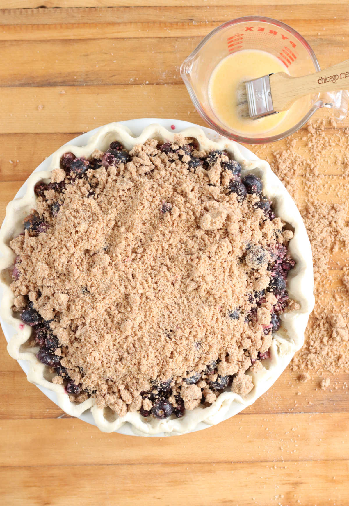 Blueberry pie with crumb topping on wooden cutting board, egg wash and pastry brush in glass measuring cup.