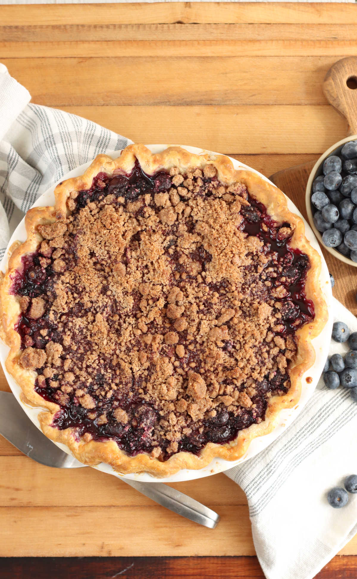 Blueberry pie crumble topping on butcher block, metal pie server and fresh blueberries.