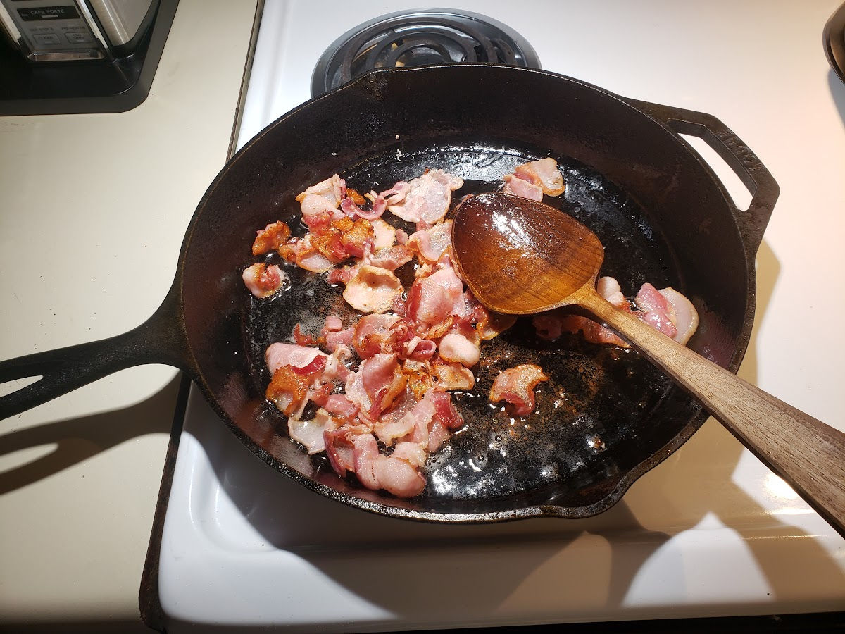 Browning bacon in large cast iron skillet, wooden spoon in pan.