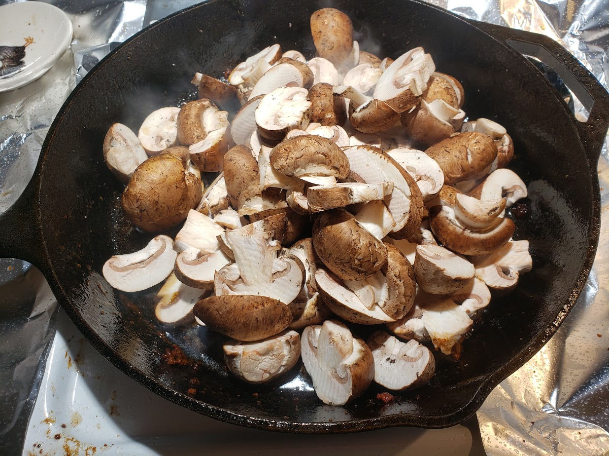 Browning mushrooms in large cast iron skillet on white kitchen stove.