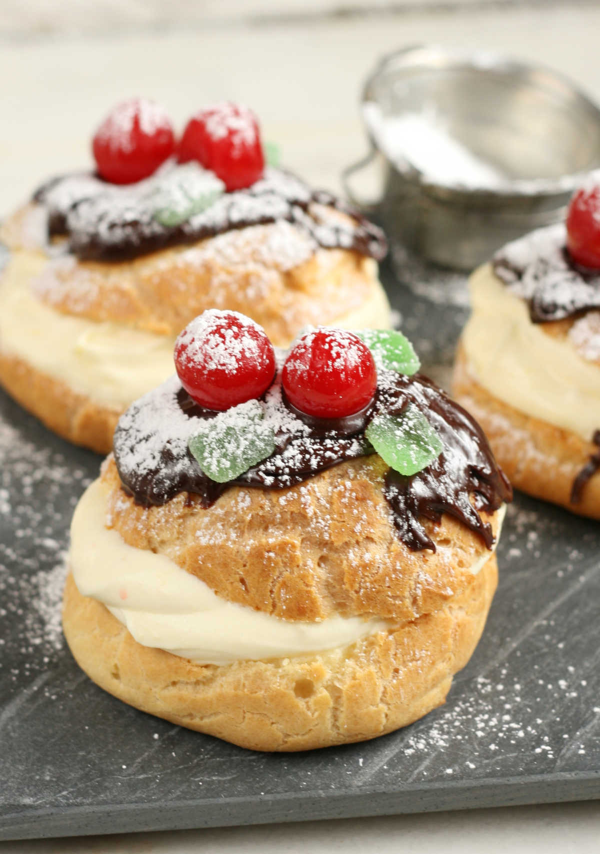 Cream puffs with pastry cream, chocolate ganache, maraschino cherries, green leaf candies dusted with powdered sugar.