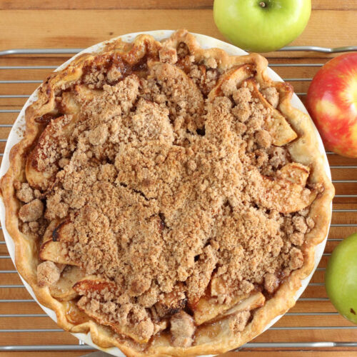 Crumb apple pie on wooden cutting board, red and green apples around.