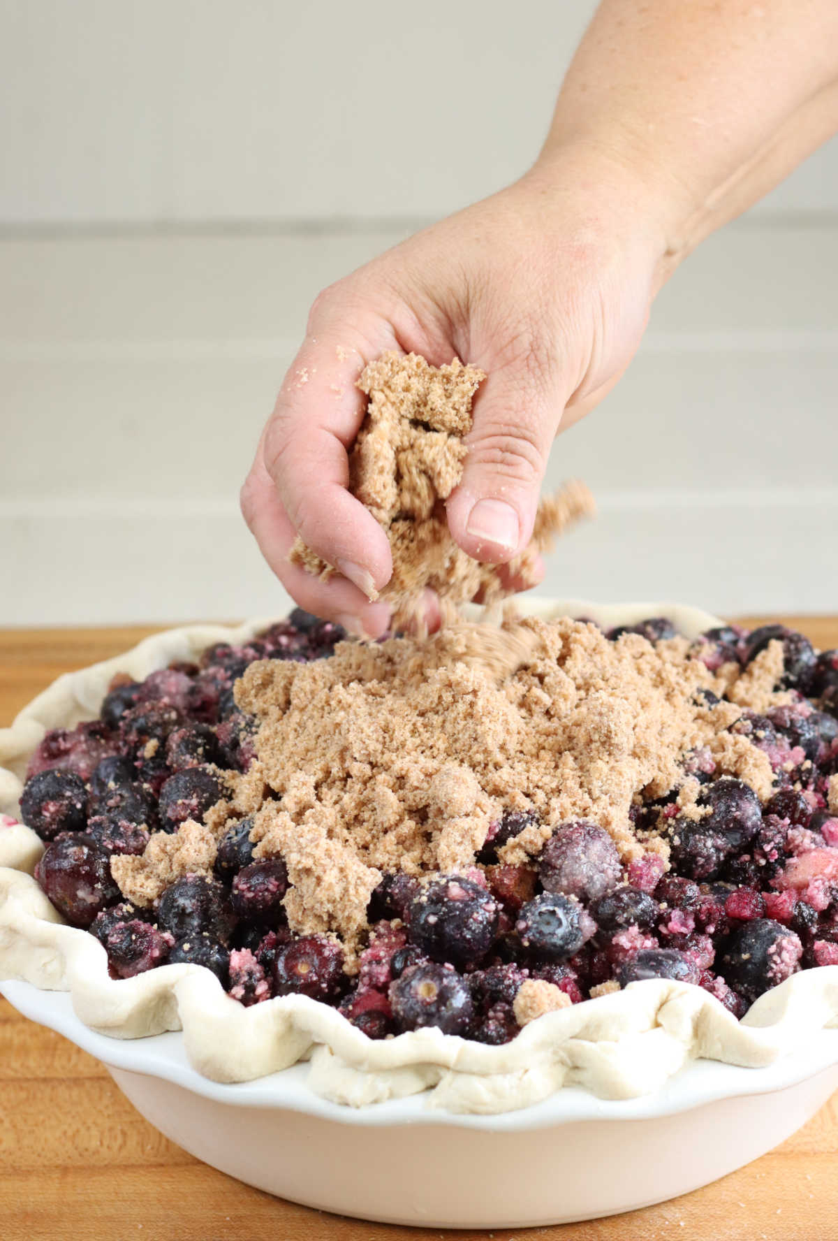 Hand pouring crumb topping on blueberry pie in white pie dish on butcher block.