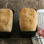 Two loaves of sourdough bread in cast iron loaf pans on metal baking rack.