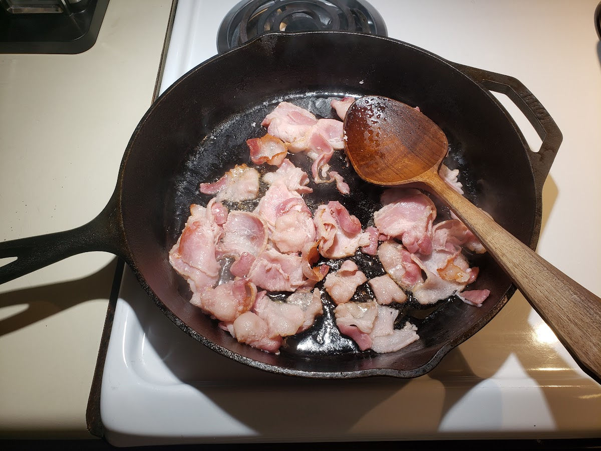 Browning bacon in large cast iron skillet on white kitchen stove.