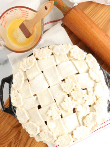 Unbaked pie with lattice crust and leaves on wooden cutting board, egg wash with pastry brush.