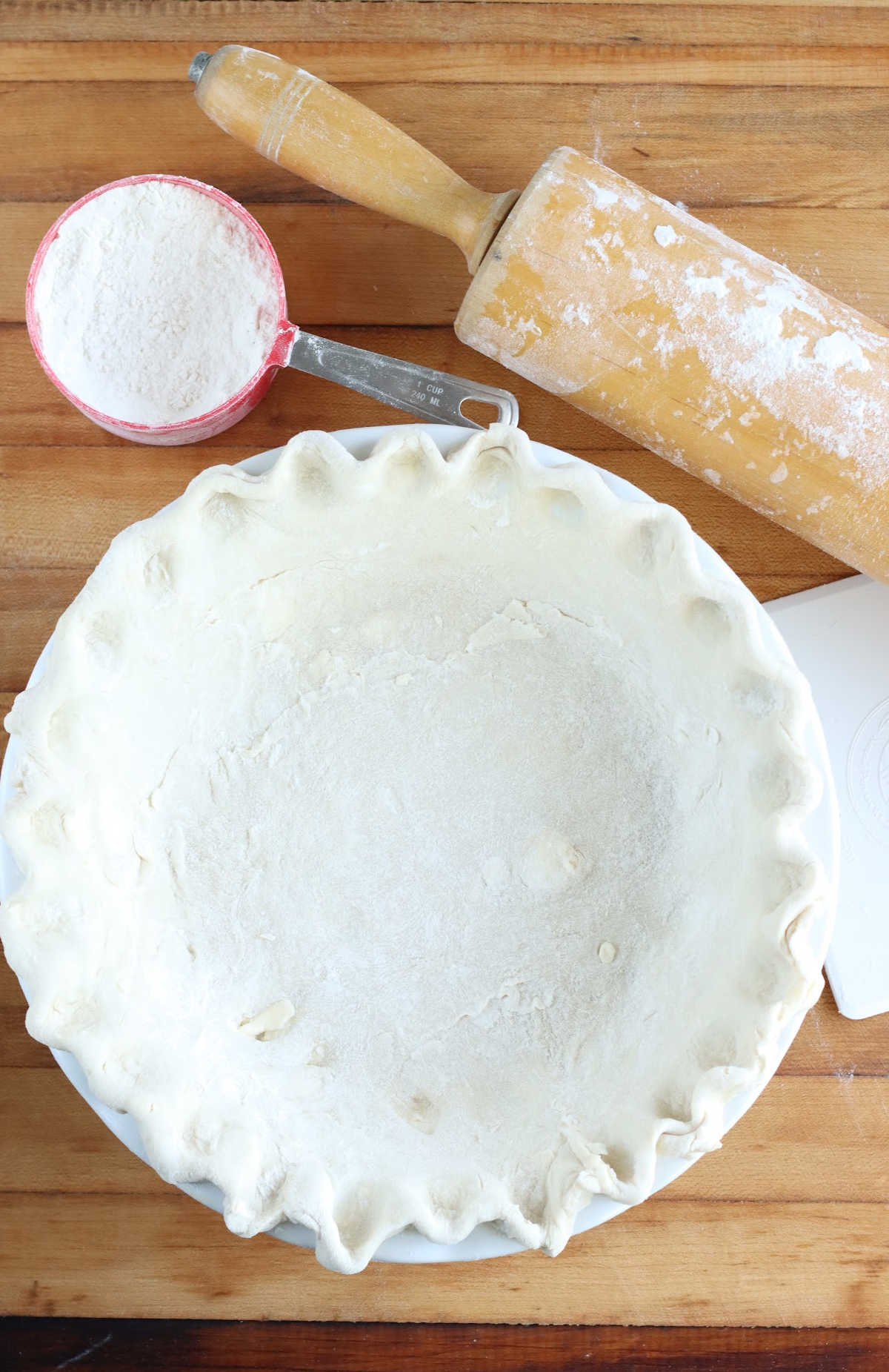 Unbaked pie shell in white pie dish on butcher block, wooden rolling pin, red measuring cup with flour.