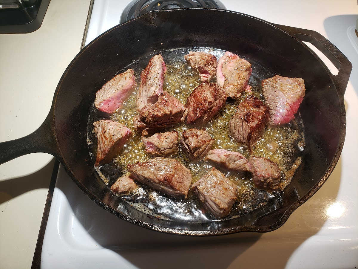 Stew beef browning in large cast iron skillet on white kitchen stove.