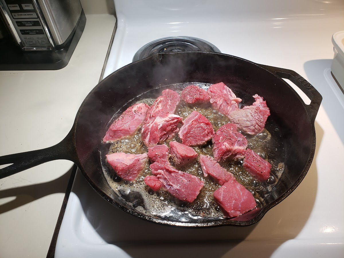 Browning stew beef in large cast iron skillet on white kitchen stove.