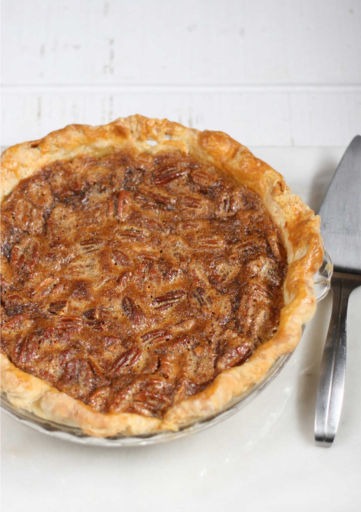 Close up of pecan pie with golden pie crust on white marble, metal pie server to right.