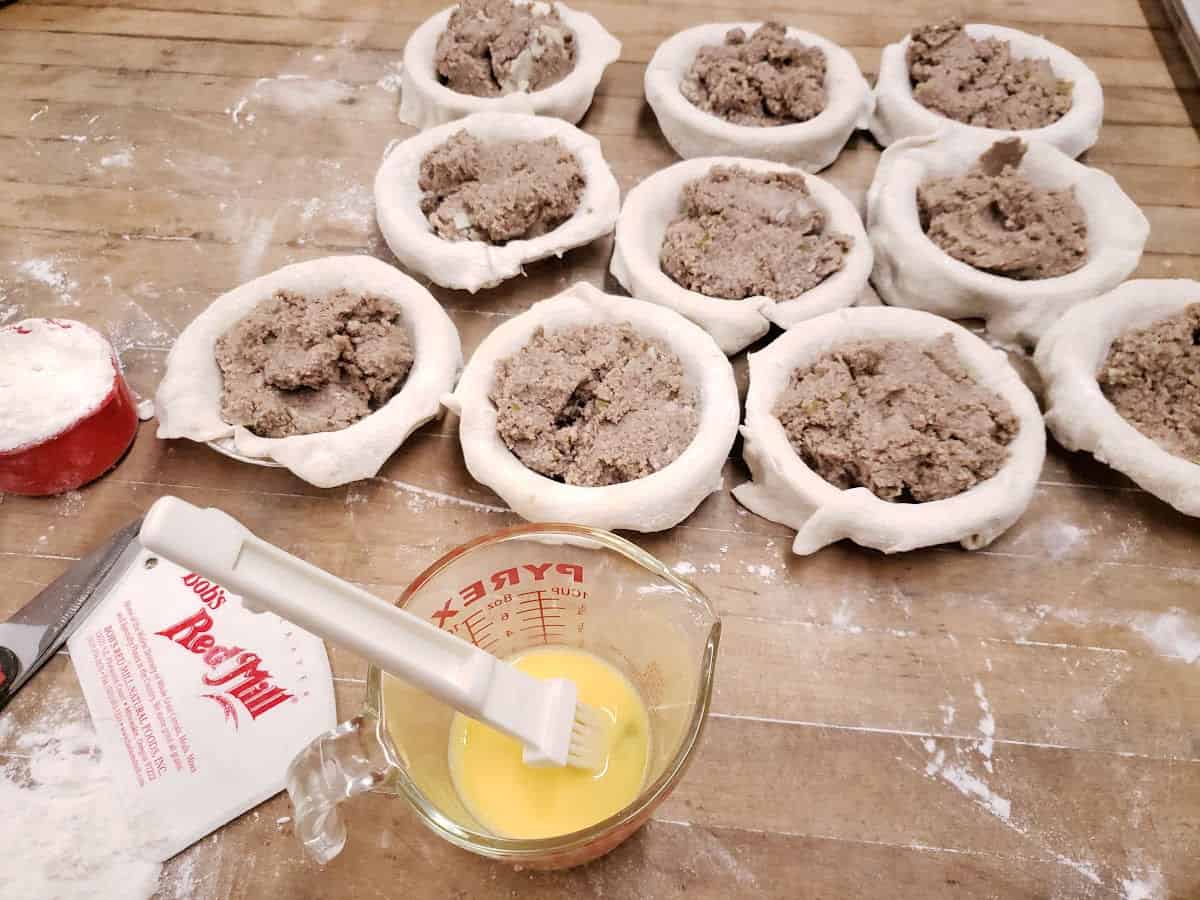 Small meat pies on butcher block before top crust is put on, egg wash in glass measuring cup.