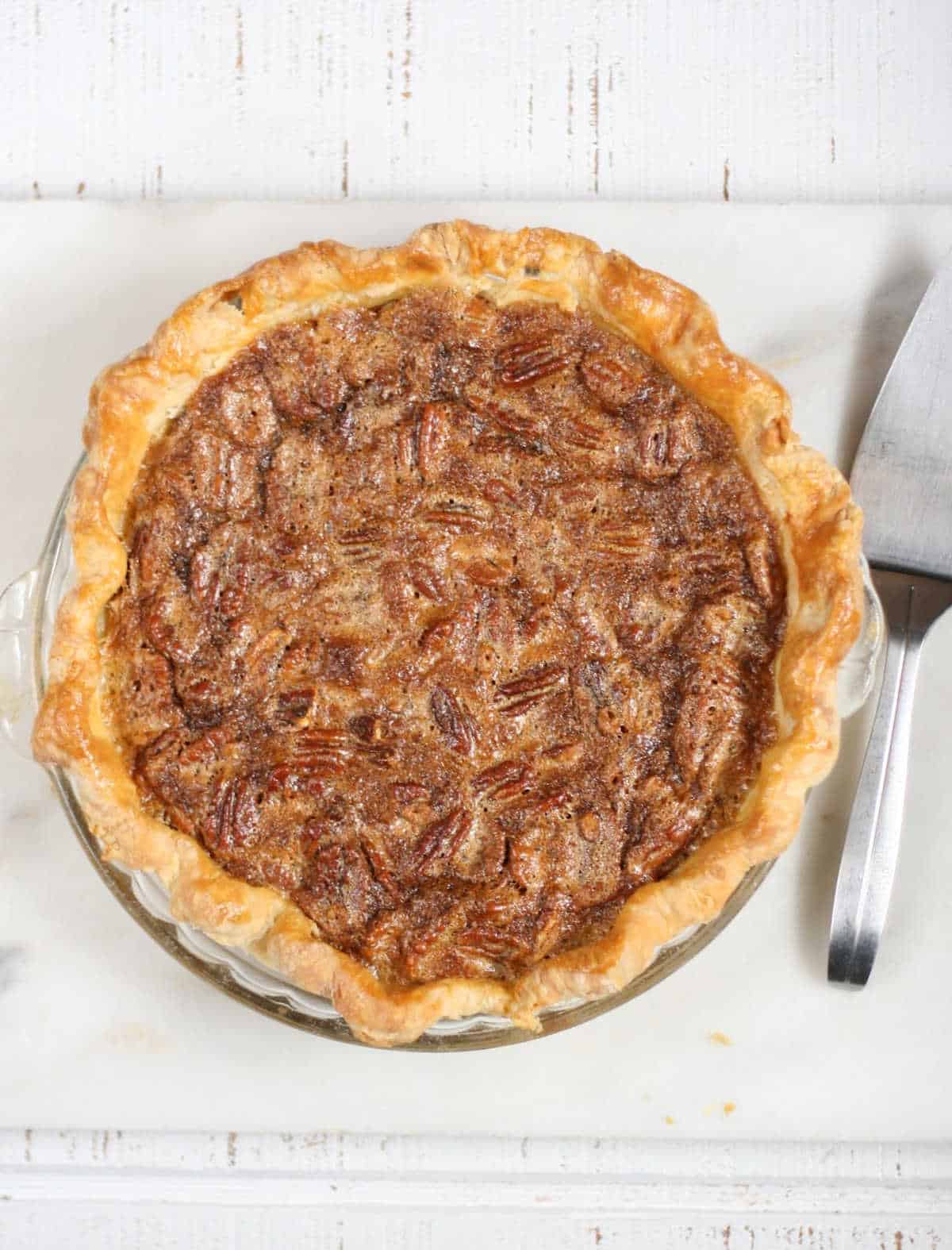Pecan pie with golden browned crust on white reclaimed wood, metal pie server to right.