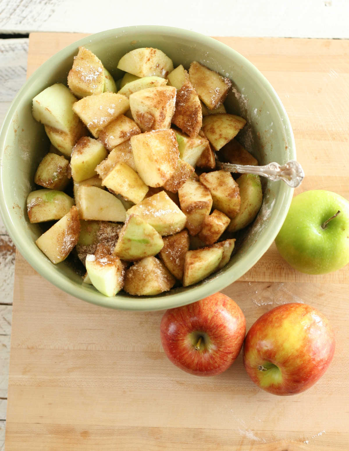Apple slices in pale green bowl on wooden cutting board, green and red apples around.