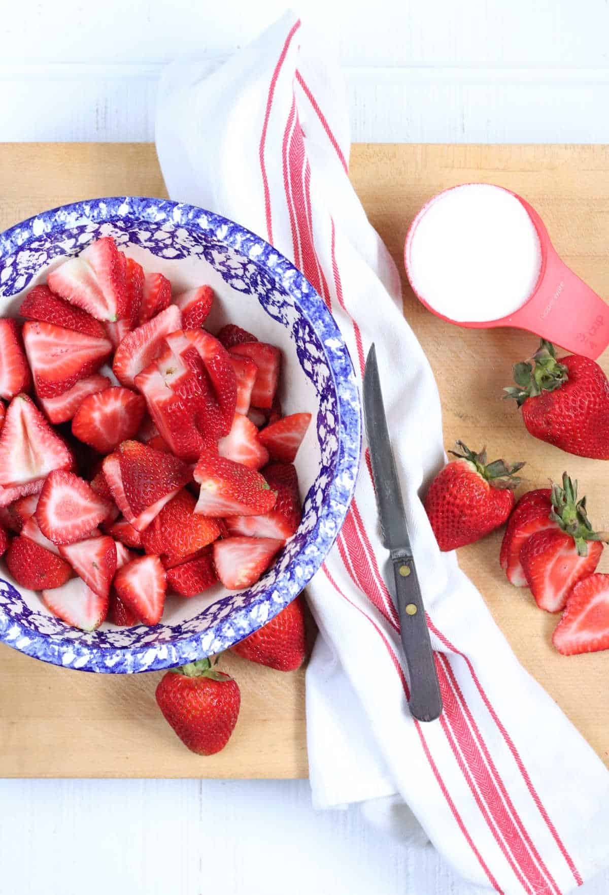 Blue speckled rim bowl with strawberry slices on wooden cutting board with knife and white/red striped kitchen towel.