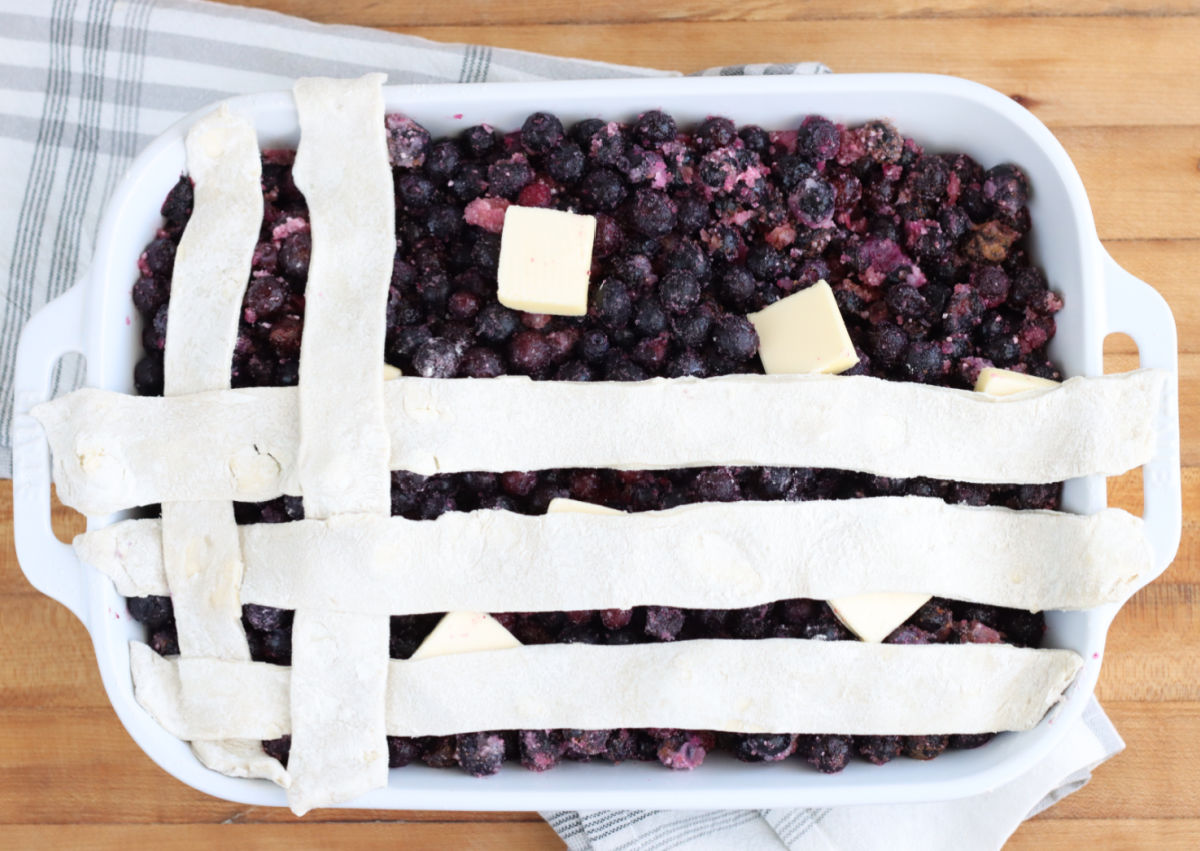 Putting lattice pie crust on blueberry cobbler in white rectangle baking dish on wooden cutting board.