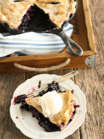 Blueberry pie in cast iron skillet, slice on small white plate.
