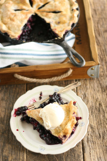 Blueberry pie in cast iron skillet, slice on small white plate.