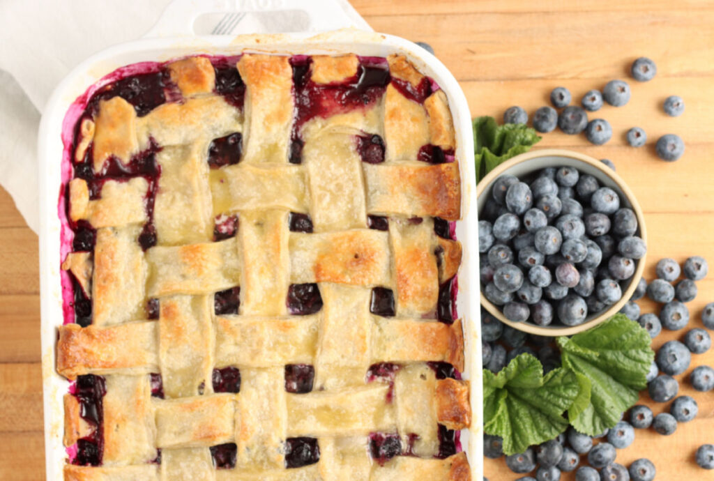 Cobbler with lattice pie crust in white rectangle baking dish on wooden cutting board, blueberries in small bowl to side.