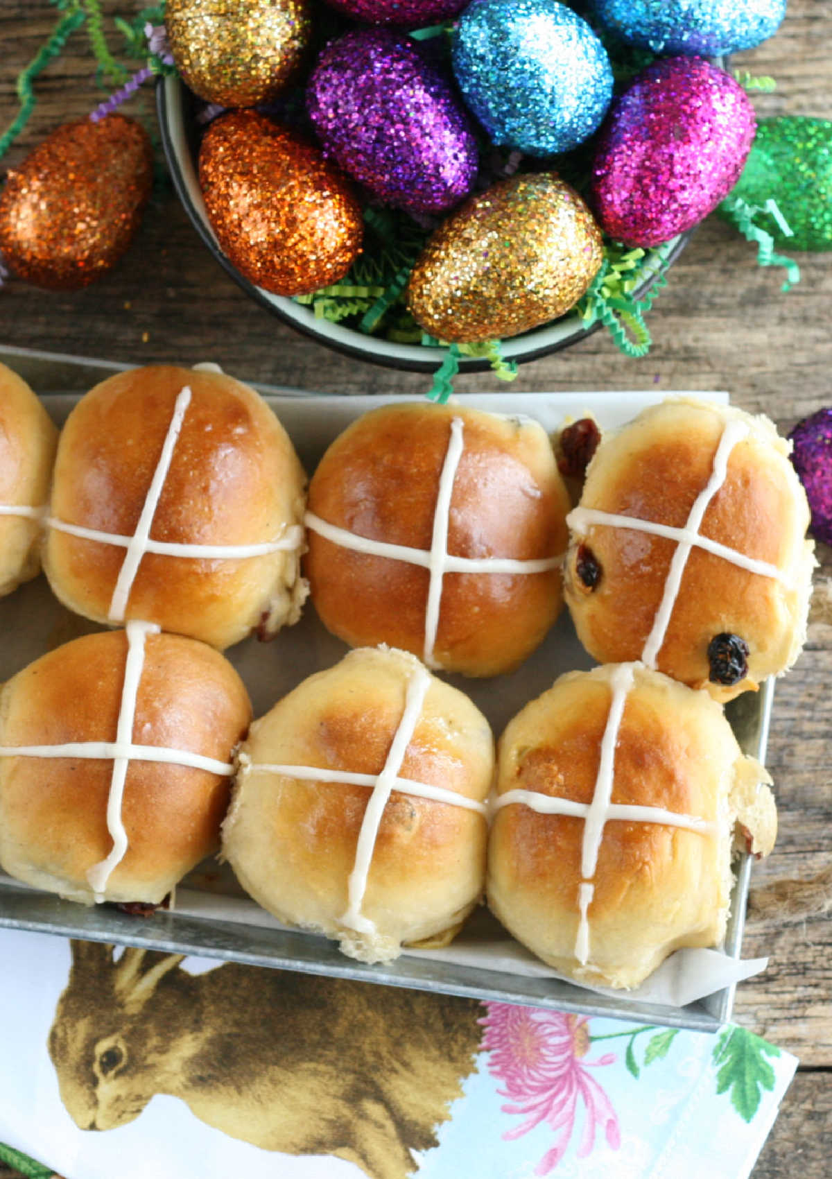 Hot cross buns with icing crosses, cranberries, colorful glittered eggs in background.