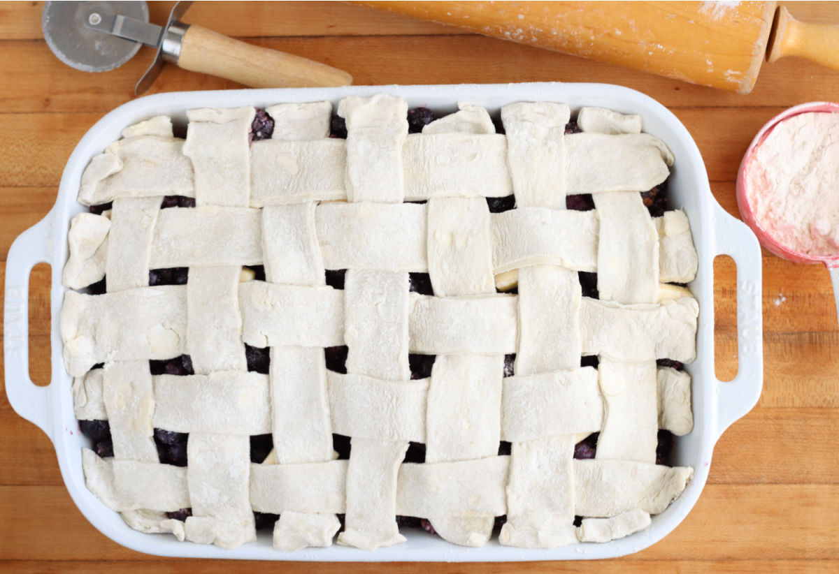 Unbaked lattice crust on cobbler in white rectangle baking dish on wooden cutting board.