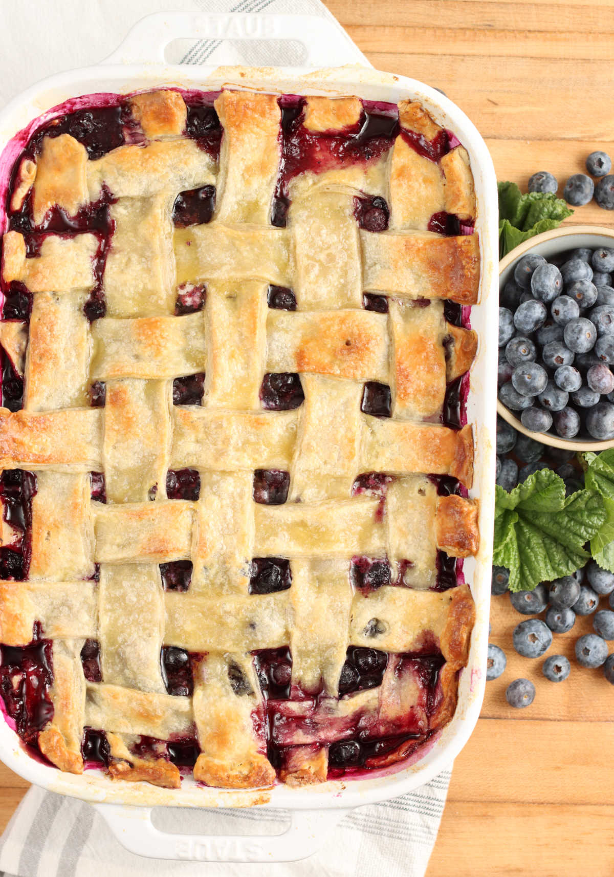 Blueberry cobbler with pie crust weaved in white rectangle baking dish on wooden cutting board, loose blueberries around.